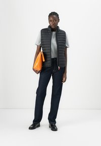 Black puffer vest over a gray t-shirt, paired with dark jeans and black shoes. Holding an orange bag with a triangular design.