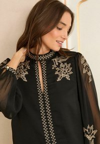Crna bluzica s prozirnim rukavima, ukrašena zlatnim cvjetnim vezom, s izrezom u obliku ključa i dekorativnim rubom duž prednje strane.