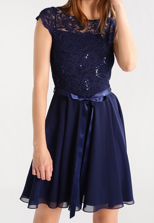 Femme portant une robe bleu marine avec de la dentelle et des paillettes sur le haut, un ruban en satin noué à la taille et une jupe en chiffon évasée.