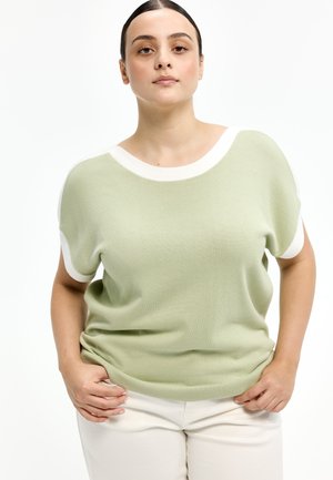 Frau mit dunklen Haaren in einem hellgrünen Stricktop mit kurzen Ärmeln und weißem Saum, stehend mit den Händen in den Taschen vor einem weißen Hintergrund.