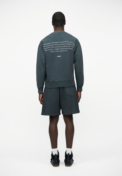 Homme portant un sweat-shirt gris foncé avec un texte imprimé blanc dans le dos, assorti à un short, des chaussettes blanches et des baskets montantes noires.