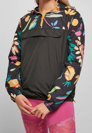 Le modèle porte une veste noire à enfiler, avec des manches fleuries et colorées et une fermeture éclair jaune, assortie à un legging rose vif.