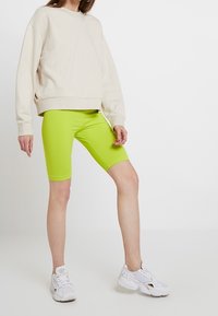 Felpa beige con orlo arrotondato, abbinata a pantaloni da ciclismo verde lime brillante. Scarpe da ginnastica bianche con dettagli decorativi.