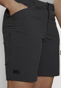 Pantalones cortos gris oscuro hechos de tela ligera. Incluyen un bolsillo con cremallera y un logo negro en la pierna izquierda. Corte entallado con detalles de costura.