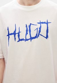 Tricou alb din bumbac, cu un logo mare, albastru, cu scris zimțat „HUGO” pe piept, cu guler rotund și mâneci scurte.