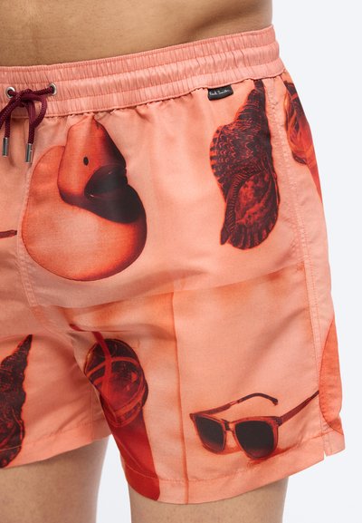 Gros plan sur un short de bain corail imprimé de canards en caoutchouc, de feuilles, de lunettes de soleil, avec un cordon noir à la taille et une petite étiquette de marque.