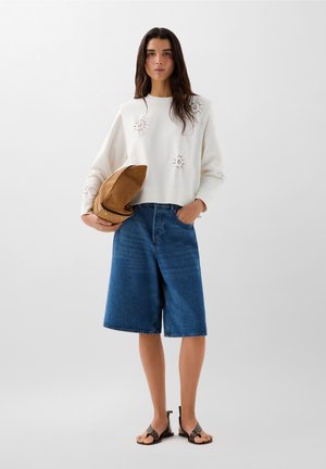Felpa crema con motivi floreali ricamati, abbinata a culotte in denim blu e sandali intrecciati marroni. Cappello nei toni della terra in mano.