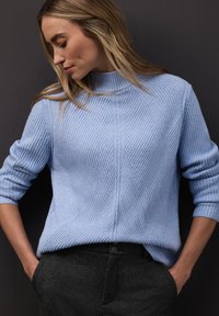 Vaaleansininen ribbineulottu paita korkealla kauluksella ja laskostetuilla olkapäillä, jossa on tekstuurillisia vinokuvioita. Tummanharmaat housut sivutaskuilla.