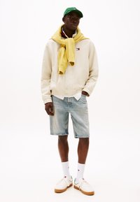 Cream bomber jacket, yellow sweater draped, blue denim shorts, white sneakers with tan soles, green cap, white socks, relaxed fit.