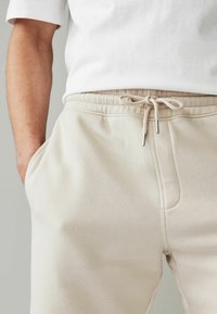 Pantalons de survêtement beige avec une taille élastique, cordon de serrage et poches latérales ; texture douce avec une finition lisse et des détails de couture minimalistes.