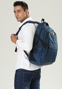 Zaino blu in nylon con spallacci imbottiti, maniglia superiore, tasche con zip e dettagli neri, portato da una persona che indossa una camicia bianca.