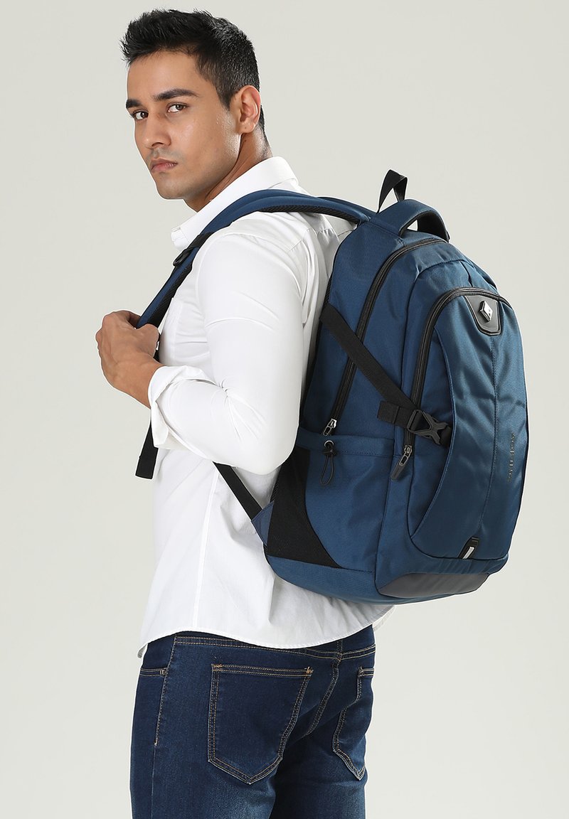 Zaino blu in nylon con spallacci imbottiti, maniglia superiore, tasche con zip e dettagli neri, portato da una persona che indossa una camicia bianca.