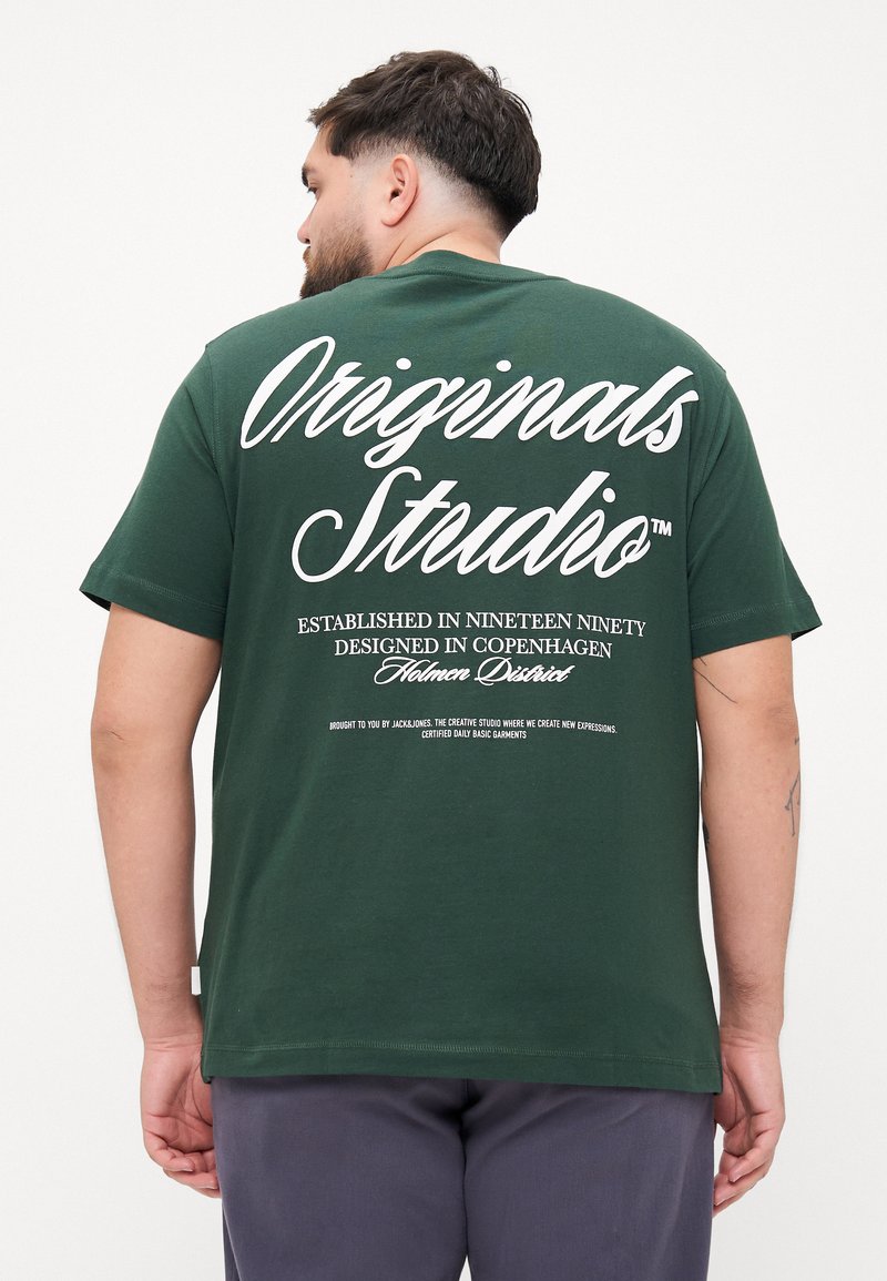 Tume roheline puuvillane T-särk valge trükitekstiga seljal. Tekst sisaldab "Originals Studio" ja täiendavaid üksikasju brändi ja disaini kohta.