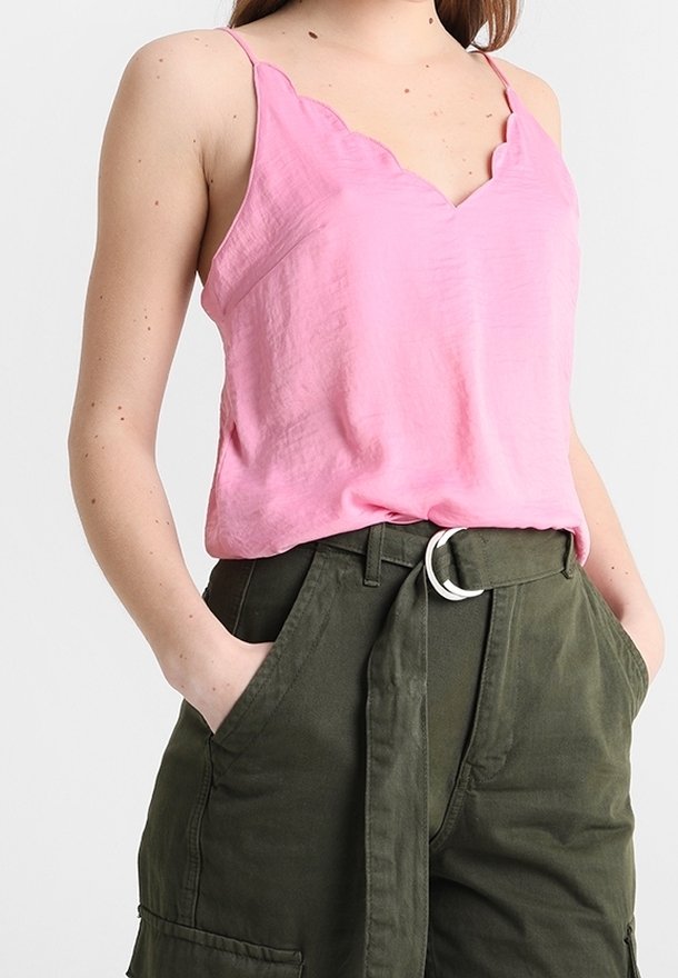 Donna che indossa un top rosa a scollatura ondulata infilato in pantaloni corti a vita alta di colore verde scuro con cintura, con le mani nelle tasche.