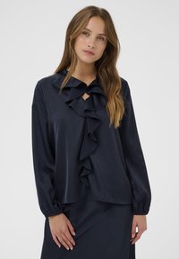 Marineblouse met een gerimpelde halslijn en voorkant, lange mouwen met elastische manchetten, en een gladde textuur. Keyhole-detail bij de hals.