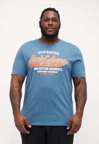 Gaiši zils kokvilnas T-krekls ar īsām piedurknēm. Iezīmēts ar lielu baltu un oranžu grafisku attēlu, tostarp tekstu "Superior Jack&Jones."