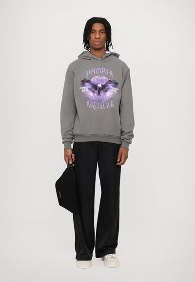 Felpa grigia con un design grafico viola raffigurante un uccello e una luna, abbinata a pantaloni neri e sneakers chiare, che tiene una borsa nera.
