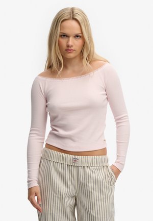 Ung kvinde med langt blondt hår iført en lyserød off-shoulder bluse med lange ærmer og beige bukser med striber, med hænderne i lommerne.