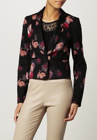 Blazer cintré noir avec motif floral rose, rouge et violet, porté sur un haut en dentelle noire, associé à un pantalon en cuir beige.
