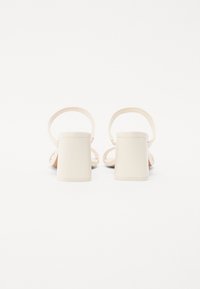 Sandalias de tacón alto beige con tacón cuadrado, tira delgada en el tobillo y textura de superficie suave. Diseño minimalista con color neutro.