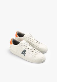 Cremefarbene Wildledersneaker mit einem orangefarbenen Fersenelement, ausgestattet mit einem schwarzen Totenkopf- und Kreuzbones-Design, flachen Schnüren und einer strukturierten Sohle.