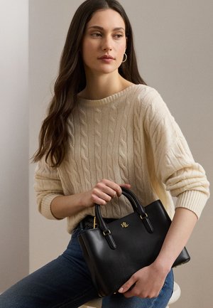 Γυναίκα με μακριά σκούρα μαλλιά που φοράει ένα κρεμ πλεκτό πουλόβερ με σχέδιο κόμπων και τζιν, κρατώντας μια μαύρη δερμάτινη τσάντα με χρυσά μεταλλικά μέρη.