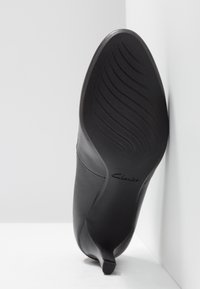Czarny but na wysokim obcasie z gładką skórzano-podobną cholewką i teksturowaną gumową podeszwą z poziomymi rowkami. Mały, spiczasty detal obcasa.