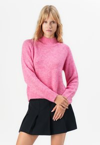 Mulher vestindo um suéter de malha rosa com gola alta e saia preta plissada, em pé contra um fundo branco com as mãos unidas.