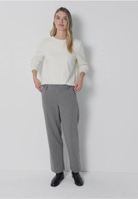 Haut blanc à manches longues associé à un pantalon rayé gris. Le pantalon a une coupe droite et des poches latérales. Le mannequin porte des bottines noires.