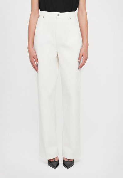 Pantalons blancs taille haute avec un design à jambes droites, dotés d'une fermeture à bouton et à zip à l'avant, et d'une texture lisse. Portés avec des talons noirs.