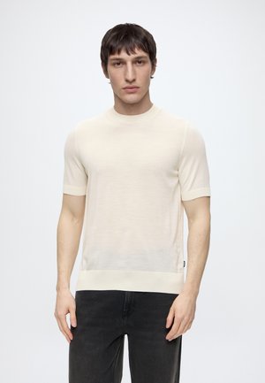 Giovane uomo con capelli corti e scuri che indossa un maglione crema a maniche corte e pantaloni neri, in piedi davanti a uno sfondo bianco semplice.