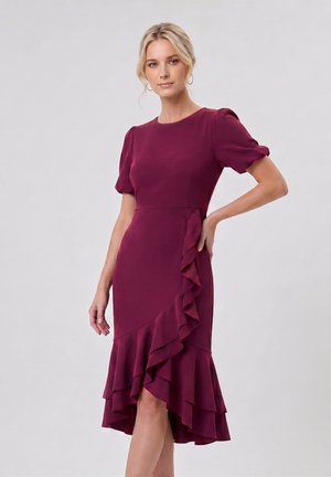 Femme aux cheveux blonds portant une robe violette avec des manches bouffantes et des détails à volants, posant avec une main sur la hanche sur un fond uni.