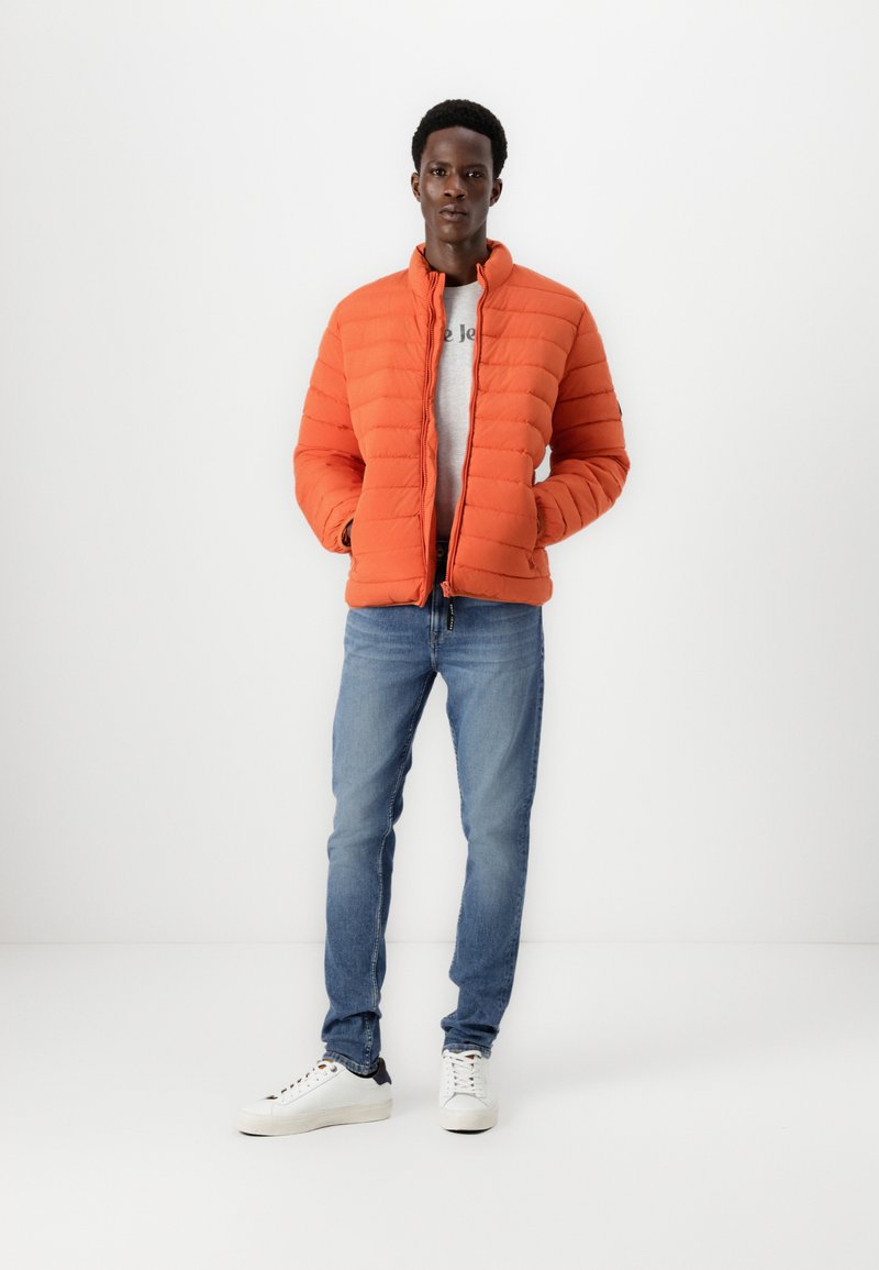 Joven vestido con una chaqueta acolchada naranja sobre una camisa gris, jeans azules y zapatillas blancas, de pie contra un fondo blanco liso.