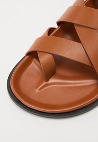 Sandale en cuir marron avec des lanières croisées, une semelle intérieure lisse et une semelle en caoutchouc noir. Le design est minimaliste et contemporain.