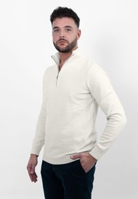 Lichtbeige ritsvest met hoge kraag, gemaakt van zachte stof. Heeft ribgebreide manchetten en zoom, gedragen over donkere broeken.
