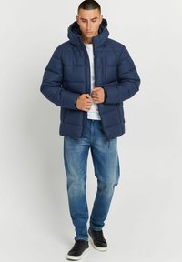 Marineblauer Pufferjacke mit strukturiertem Finish, hohem Kragen und Kapuze. Kombiniert mit hellblauen Jeans und schwarzen Sneakers, steht aufrecht.