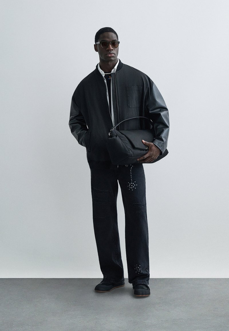 Black oversized bomber jacket with leather sleeves, paired with dark denim pants featuring studded accents and a black handbag.