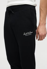 Μαύρη φόρμα με τσέπες στο πλάι και λευκό λογότυπο «Jack & Jones Archive Selection», φορεμένη με λευκό πουκάμισο, που δείχνει μέρος του δεξιού χεριού.