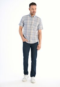 Camicia a maniche corte con bottoni, motivo a quadrati blu e beige, abbinata a jeans scuri e sneakers bianche. Il modello sta in piedi con le mani nelle tasche.