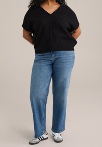 Černý texturovaný pletený kardigan s výstřihem do V a krátkými rukávy, kombinovaný se světlomodrými volnými džíny a černobílými teniskami.