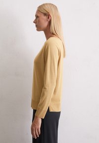 Senfgelbes Strickpullover mit langen Ärmeln, rundem Halsausschnitt, seitlichen Schlitzen und lockerer Passform, kombiniert mit dunklen Unterteilen. Geschmeidige Textur.