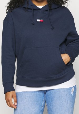 Person wearing a navy blue Tommy Jeans hoodie with front pocket and light blue ripped jeans, hand in pocket and curly hair visible.