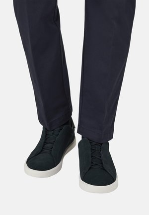 Persona con scarpe sportive nere con suole bianche e pantaloni blu scuro in piedi su sfondo bianco.