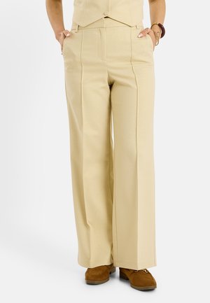 Femme portant un pantalon beige à jambes larges, les mains dans les poches et des chaussures en daim marron, cadrée à la taille et aux pieds.