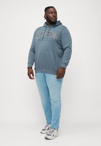 Blue cotton hoodie with a large logo, front kangaroo pocket, ribbed cuffs, paired with light blue denim jeans and gray sneakers.