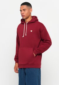 Man wearing a maroon hoodie with white drawstrings and small bee logo, standing with one hand in the front pocket, blue jeans.