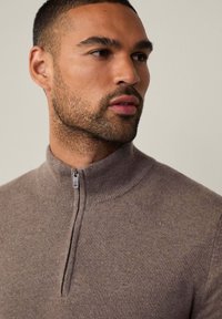 Maglione con zip in morbido tessuto marrone testurizzato. Presenta un colletto a coste e sottili dettagli di cucitura vicino alla cerniera.