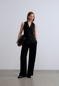 Μαύρο αμάνικο τοπ με V-λαιμόκοψη, συνδυασμένο με μαύρο παντελόνι φαρδιάς γραμμής. Τα αξεσουάρ περιλαμβάνουν μια μεγάλη μαύρη τσάντα φάκελο και διακριτικά σκουλαρίκια.