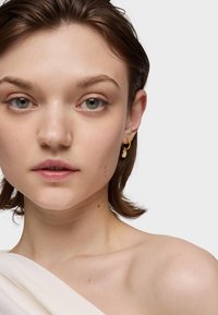 Goldene Creolen mit einem kleinen blauen Akzent, getragen von einer Person mit kurzen Haaren. Weiches, cremefarbenes Material fällt über eine Schulter.