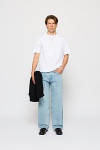 Witte ronde hals t-shirt, lichtblauwe spijkerbroek met wijde pijpen, houdt een zwarte geribbelde trui vast. Donkere schoenen maken de casual look compleet.